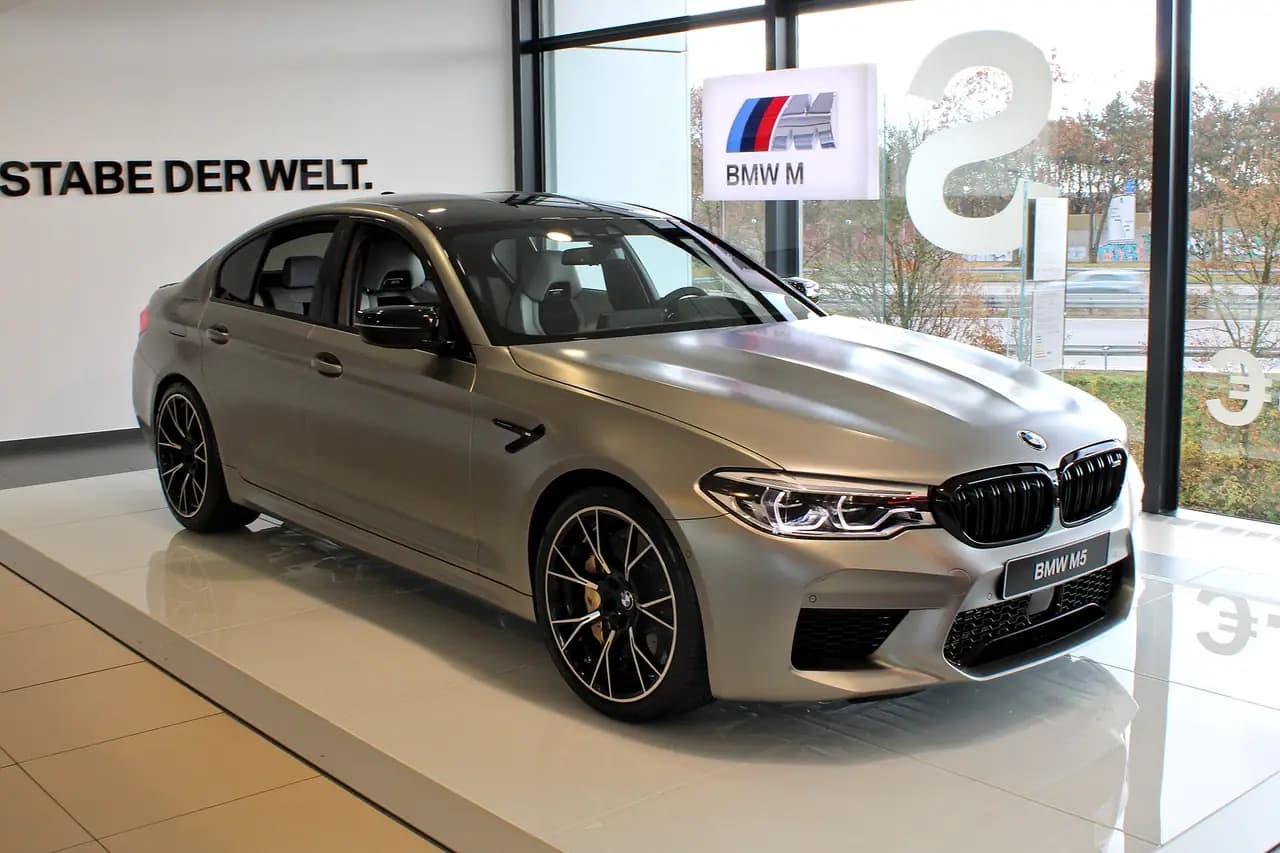 Matte silver BMW M5 Competition (F90, pre-LCI) sedan in a BMW showroom, front three-quarter view